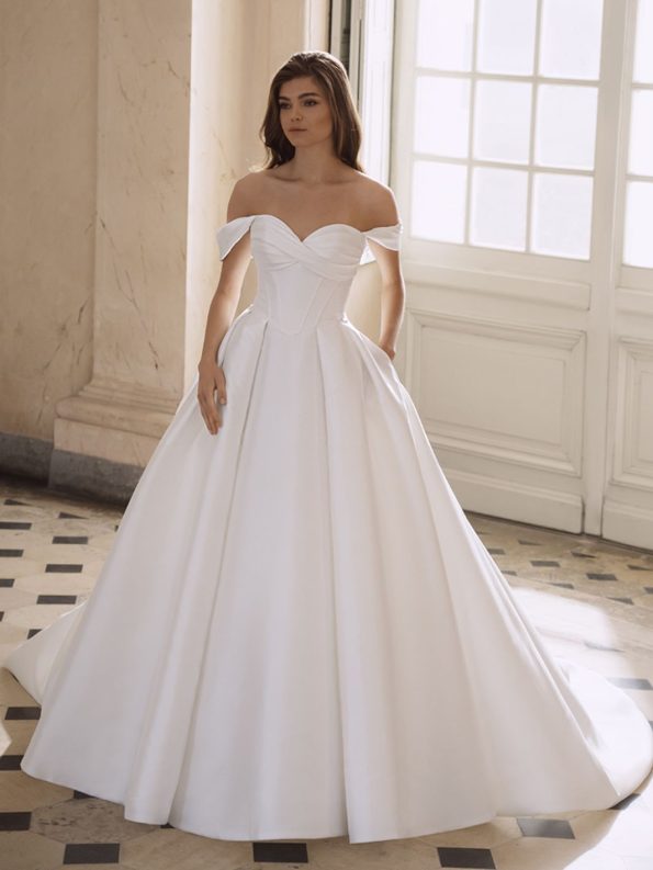 A woman stands indoors wearing the Freya by Etoile off-the-shoulder white bridal gown with a fitted bodice, full skirt, and pockets where her hands rest.