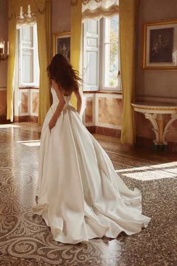 A woman in the 88377/IVA by Justin Alexander wedding dress stands in an elegant, sunlit room with large windows, yellow curtains, and ornate flooring.