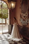 A woman in a white, sleeveless 88377/IVA by Justin Alexander wedding dress stands indoors beside a decorated mirror, with one hand resting on a marble surface.