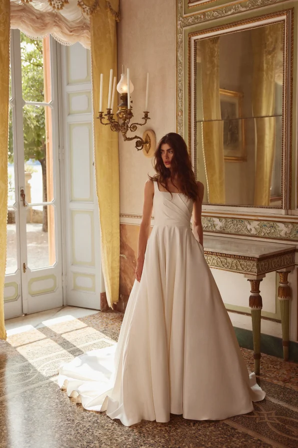 A woman in the 88377/IVA by Justin Alexander, a white floor-length wedding dress, stands indoors near a tall window as sunlight streams into an elegant, ornate room.