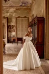 A woman wears the 88436/KELSEY by Justin Alexander—a strapless, full-skirted gown—standing indoors with hands on hips, gazing to the side in an ornate, classic room.