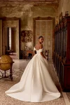 A woman wears the 88436/KELSEY by Justin Alexander—a strapless, full-skirted gown—standing indoors with hands on hips, gazing to the side in an ornate, classic room.