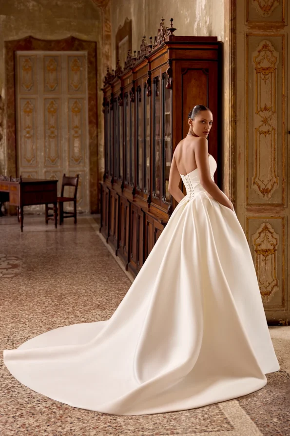A woman in the 88436/KELSEY by Justin Alexander strapless bridal gown, featuring a long train, stands in an ornate hallway and glances back over her shoulder.