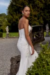 A woman in a strapless white KAI/88439 by Justin Alexander wedding dress stands outdoors near stone steps and a table set for an event.