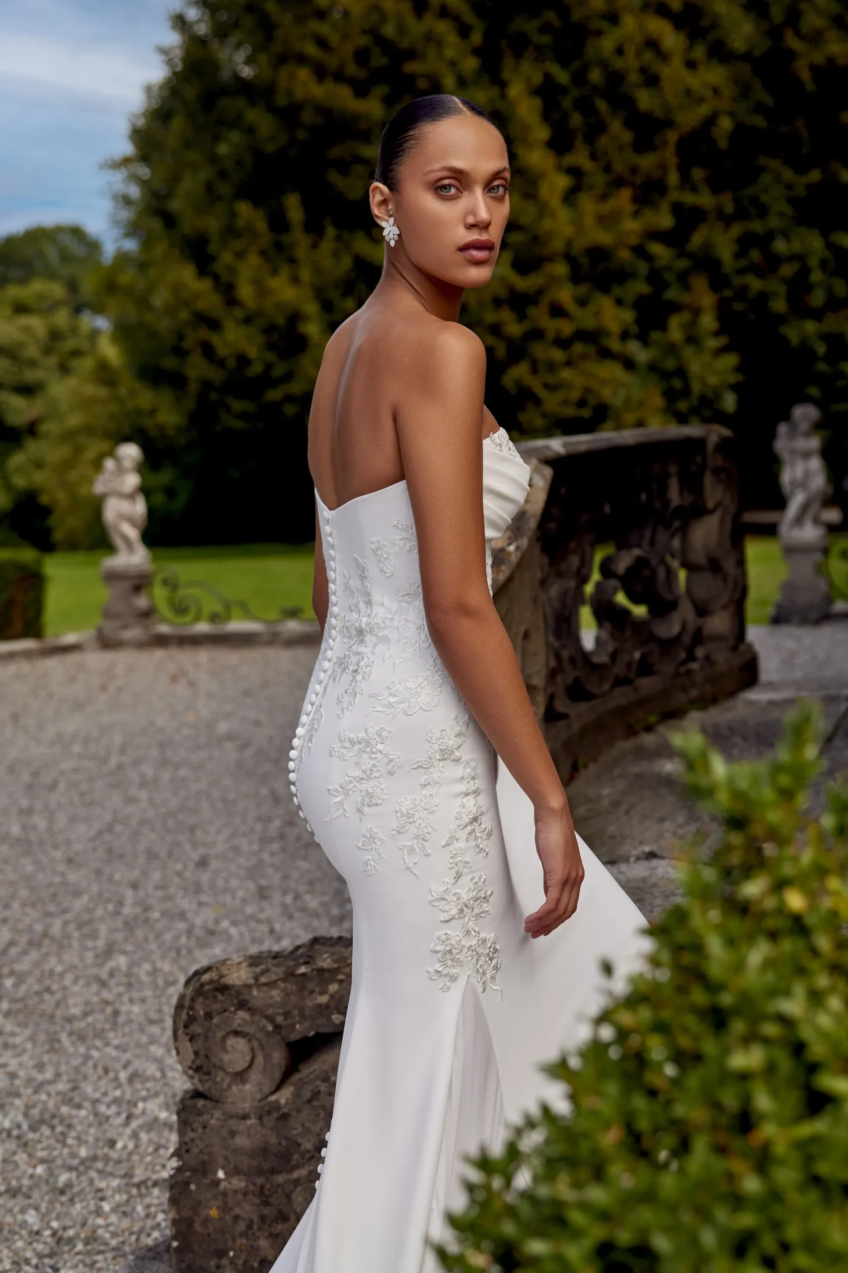 A woman in the KAI/88439 by Justin Alexander, a strapless white wedding dress with floral embroidery, stands outdoors near a stone railing and looks over her shoulder. A woman in the KAI/88439 by Justin Alexander, a strapless white wedding dress with floral embroidery, stands outdoors near a stone railing and looks over her shoulder.