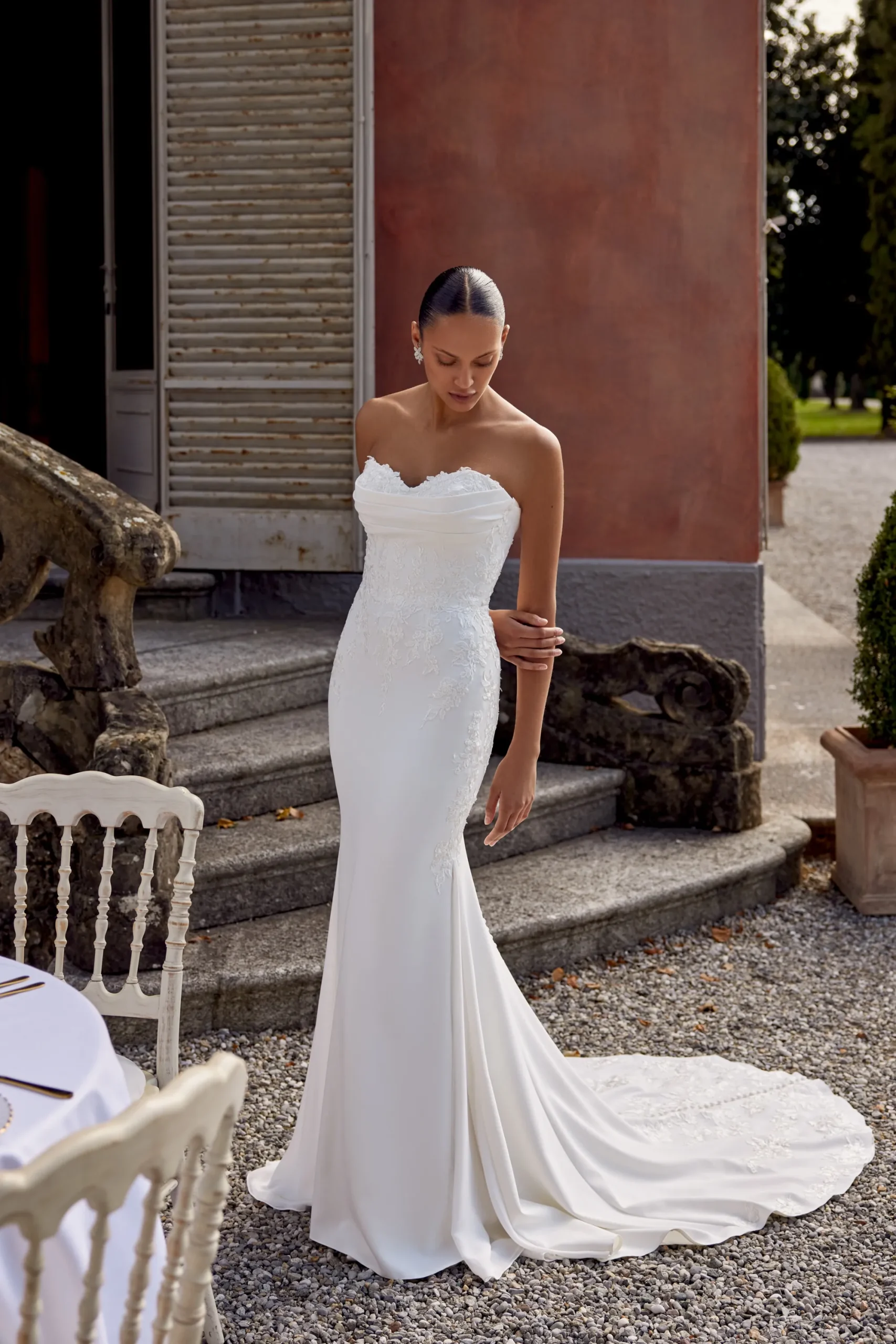 A woman in a strapless white KAI/88439 by Justin Alexander wedding dress stands outdoors near stone steps and a table set for an event. A woman in a strapless white KAI/88439 by Justin Alexander wedding dress stands outdoors near stone steps and a table set for an event.