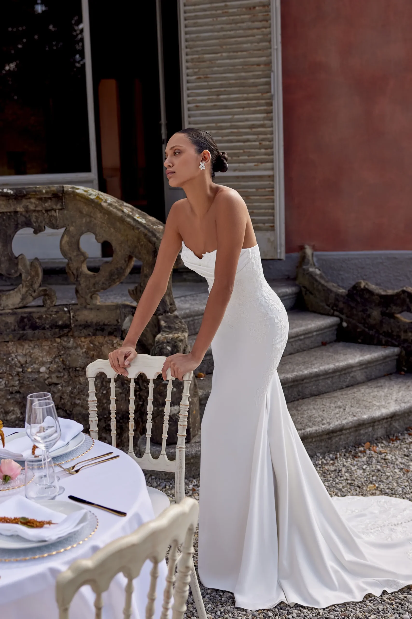 A woman in a white KAI/88439 by Justin Alexander wedding dress stands outdoors by a formally set table near stone steps and a building. A woman in a white KAI/88439 by Justin Alexander wedding dress stands outdoors by a formally set table near stone steps and a building.