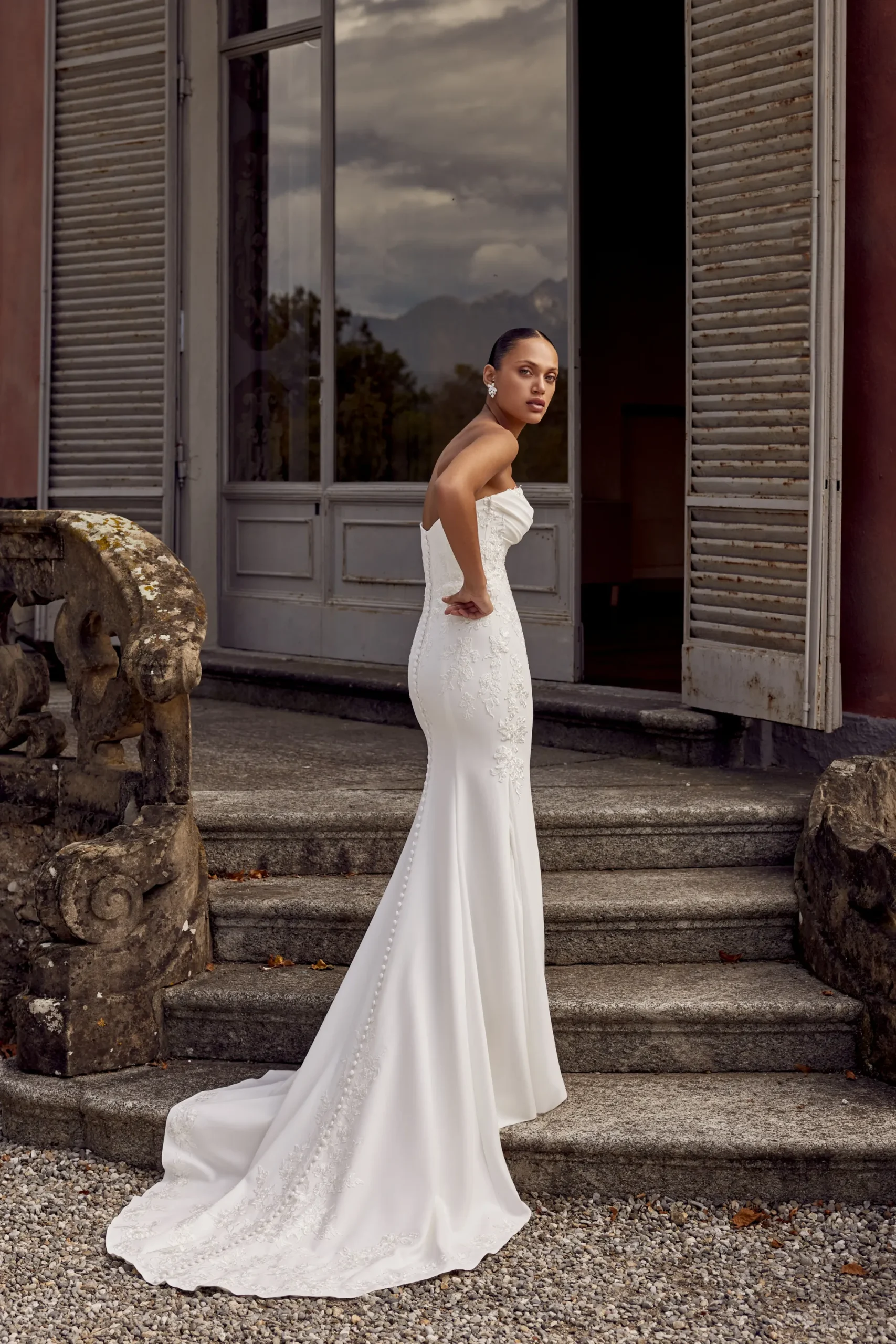 A woman in the KAI/88439 strapless white Justin Alexander wedding gown stands on stone steps outside a building with open shutters and glass doors. A woman in the KAI/88439 strapless white Justin Alexander wedding gown stands on stone steps outside a building with open shutters and glass doors.