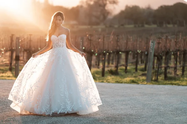 A woman in the A1252 by Allure strapless white lace wedding dress stands outdoors at sunset, holding her gown with vineyards and lush greenery in the background.
