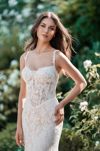 A woman stands outdoors in the A1316 by Allure, a fitted lace bridal gown with thin straps, surrounded by greenery and white flowers.