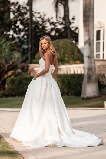 A woman in the A1350 by Allure strapless white wedding gown stands outdoors on a paved path, surrounded by greenery and palm trees, looking over her shoulder.