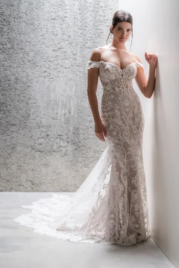A woman in the C685 by Allure, an off-shoulder lace gown with a sheer train, stands against a textured wall.