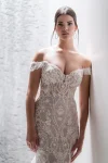 A woman in the C685 by Allure, an off-shoulder lace gown with a sheer train, stands against a textured wall.