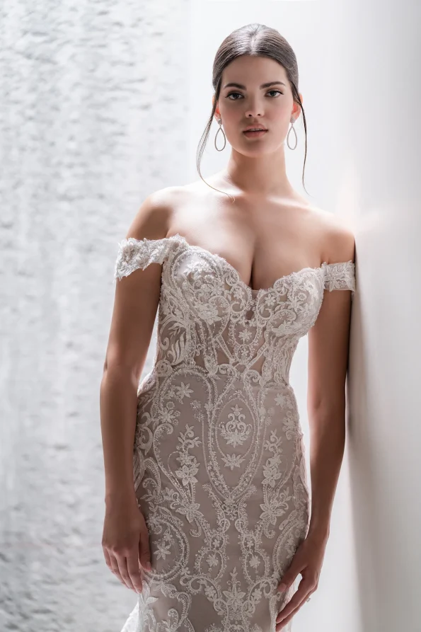 A woman stands against a light wall wearing the C685 by Allure, an off-the-shoulder, fitted wedding dress with lace embroidery and floral patterns, paired with hoop earrings.