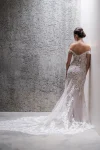 A woman in the C685 by Allure, an off-shoulder lace gown with a sheer train, stands against a textured wall.