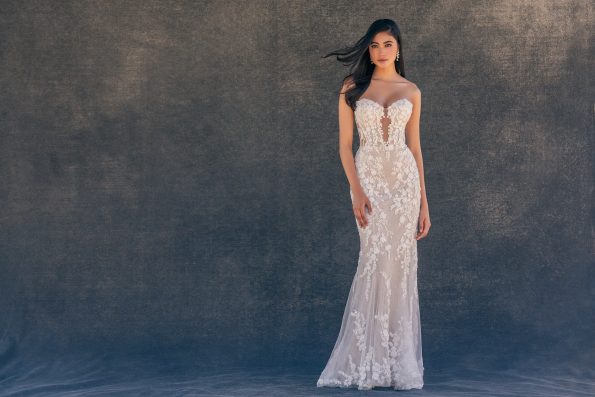 A woman in the C725 by Allure, a strapless fitted white lace gown with floral patterns, stands against a dark textured background.