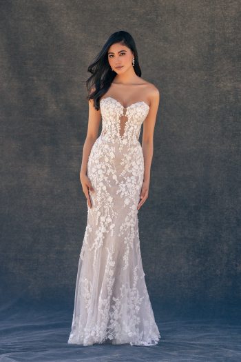 A woman stands gracefully against a plain dark background, wearing the C725 by Allure—a strapless, fitted white lace gown with floral detailing.