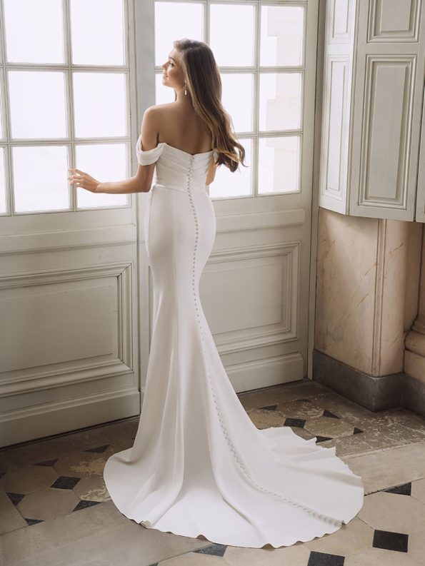 A woman stands facing a window with her back to the camera, wearing the "Fatima by Enzoani" bridal gown—an off-shoulder, fitted design featuring a button-down back and a long train.