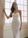 A woman in a white, off-the-shoulder Fatima by Enzoani bridal gown stands by a large window bathed in natural light, gazing slightly to the side.