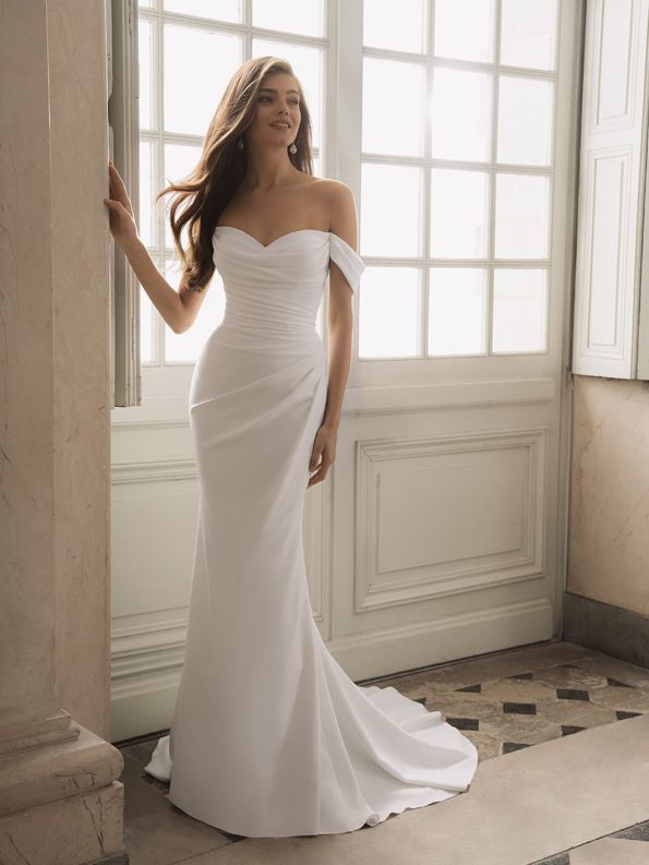 A woman in a white, off-the-shoulder Fatima by Enzoani bridal gown stands by a large window bathed in natural light, gazing slightly to the side.