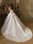 A woman stands indoors wearing the Geneva by Enzoani, a strapless white ball gown wedding dress featuring a fitted bodice, decorative embroidery, and a long train.