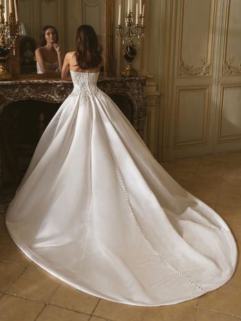 A woman wearing the Geneva by Enzoani wedding dress, featuring a long train, stands before a mirror in an ornate, classically decorated room, capturing timeless elegance.