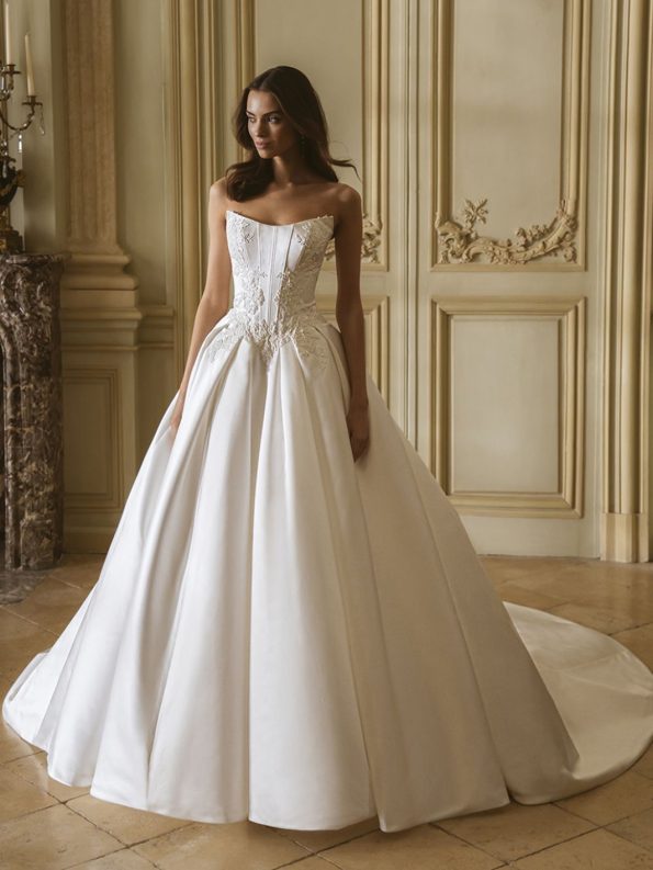 A woman stands indoors wearing the Geneva by Enzoani, a strapless white ball gown wedding dress featuring a fitted bodice, decorative embroidery, and a long train.
