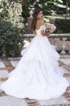 A woman in the R3865 by Allure, an off-the-shoulder white wedding dress, stands outdoors in a sunlit garden with light streaming through the trees.