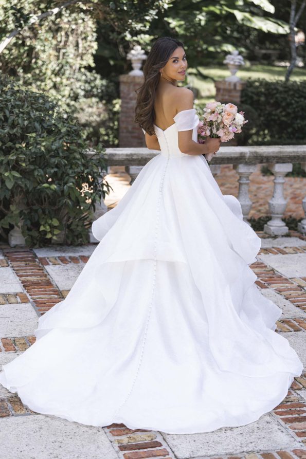 A woman in the off-the-shoulder R3865 by Allure holds a bouquet and stands outdoors on a stone patio, facing slightly backward.