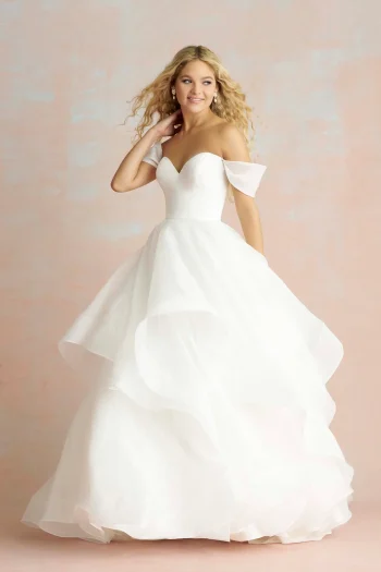 A woman in the R3865 by Allure, a white off-the-shoulder ball gown with a layered skirt, stands before a pale pink background and smiles softly.