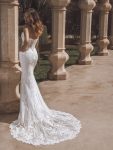 A woman stands outdoors wearing a strapless, fitted lace wedding gown with a long, flowing train and intricate embroidery details.
