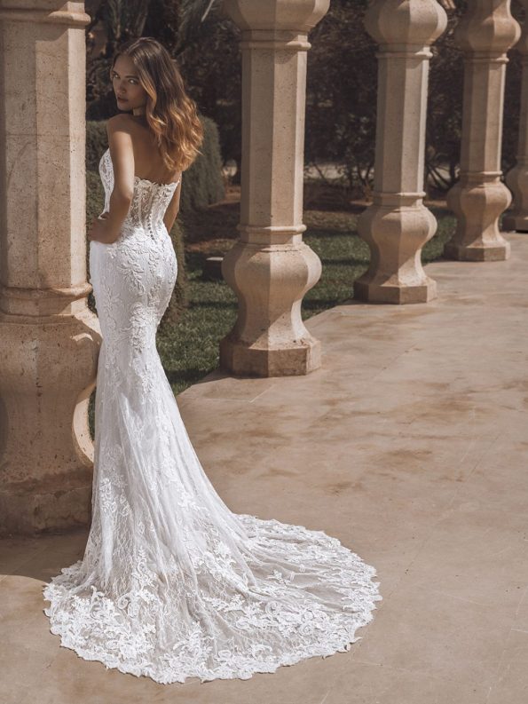 A woman wearing a strapless, form-fitting lace wedding dress with a long train stands outdoors beside stone columns.