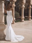 A woman stands outdoors wearing a strapless, fitted lace wedding gown with a long, flowing train and intricate embroidery details.
