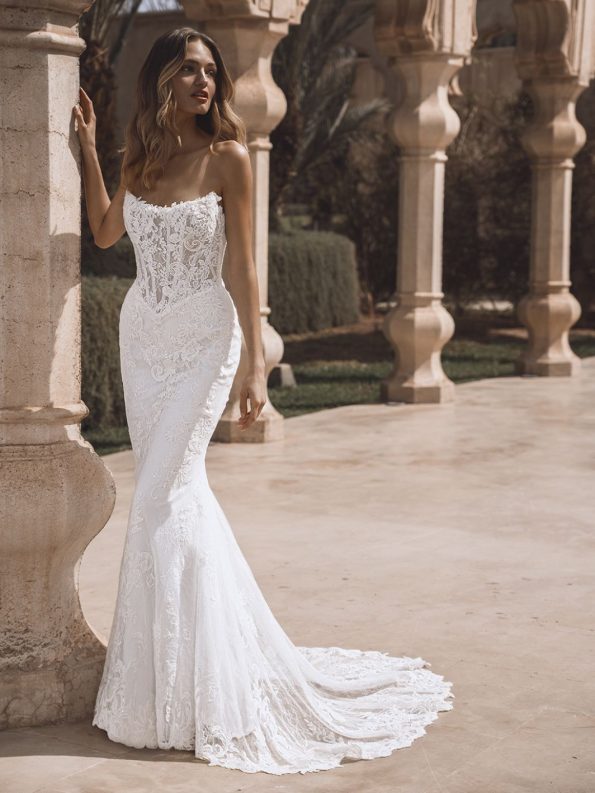 A woman wearing a fitted, strapless white lace wedding gown stands by a stone column in an outdoor setting with arches and greenery.