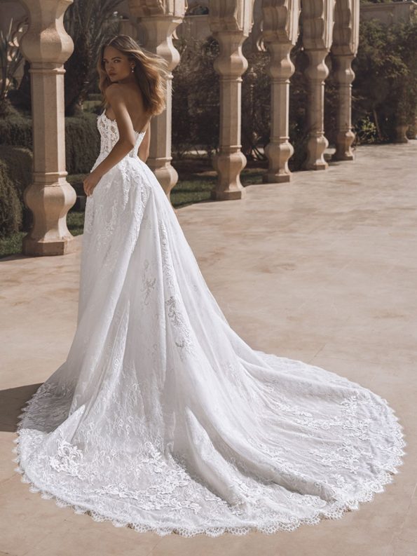 A woman in a strapless white lace wedding dress with a long train stands outdoors near stone arches, looking over her shoulder.