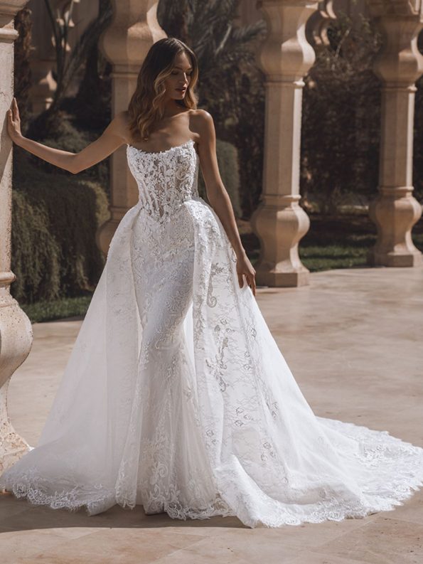 A woman stands outdoors wearing a strapless, fitted lace wedding gown with a long, flowing train and intricate embroidery details.
