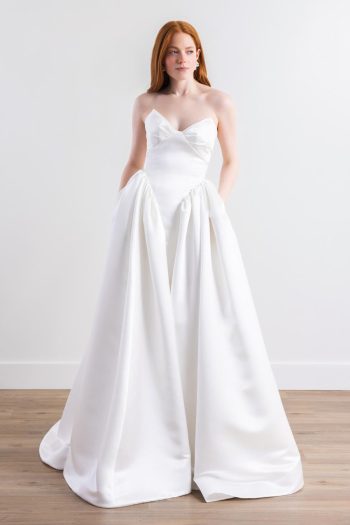A woman with long red hair stands against a plain white wall on a wooden floor, wearing the Cassia by Willowby 54319—a strapless white bridal gown with a fitted bodice and voluminous skirt.