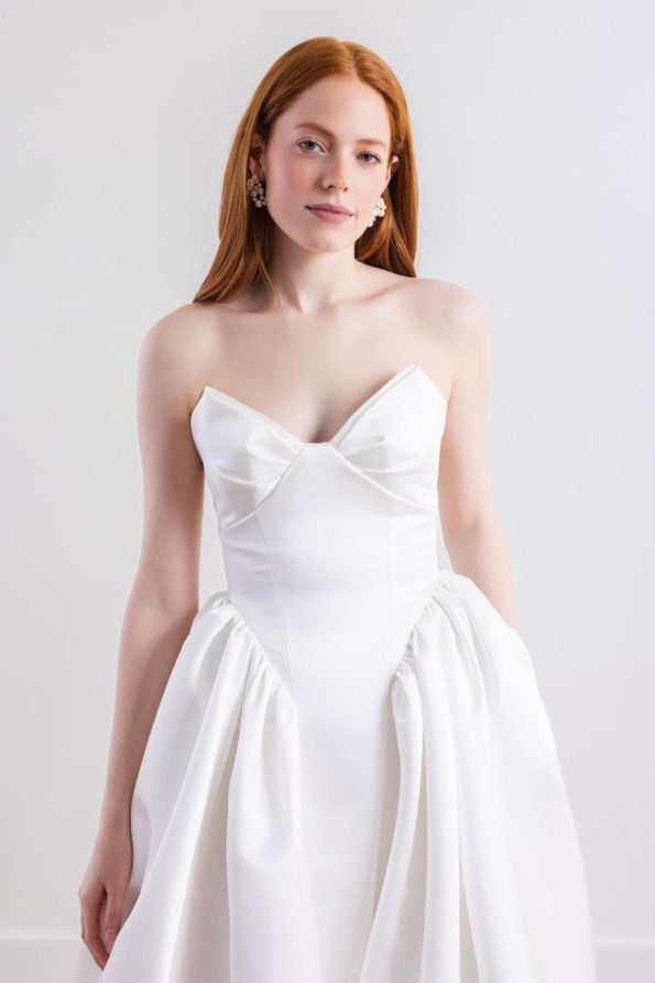 A woman with long red hair wears the Cassia by Willowby 54319, a strapless white wedding dress with a fitted bodice and gathered skirt, standing against a plain white background.