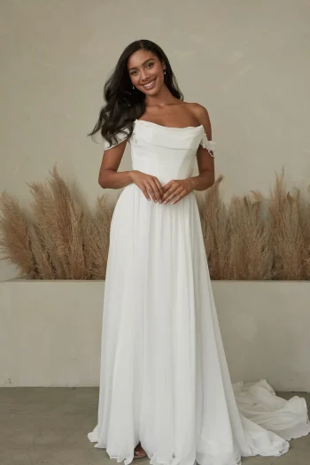 A woman stands indoors in the Bennet - Serene by Madi Lane off-the-shoulder white gown with a long, flowing skirt, as dried pampas grass decorates the background.