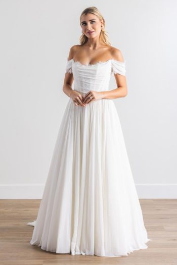 Indoors, a woman wears Calla by Wtoo 14744, an off-the-shoulder white wedding dress with a fitted bodice and flowing skirt.