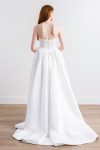A woman with long red hair stands against a plain white wall on a wooden floor, wearing the Cassia by Willowby 54319—a strapless white bridal gown with a fitted bodice and voluminous skirt.