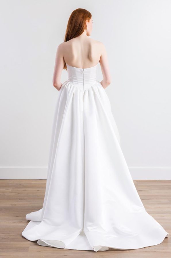 A woman with long red hair wears the Cassia by Willowby 54319 strapless white wedding gown, seen from behind on a wooden floor against a plain white wall.