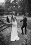 A bride in the elegant Shadow by Eva Lendel white dress, holding a bouquet, sits on a man’s lap at an outdoor event beside guests at a beautifully decorated table beneath dappled sunlight.