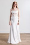 A woman wears the Rowan by Willowby 54406M, a strapless white gown with a sheer lace bodice and a long skirt, standing on a wooden floor before a plain white wall.
