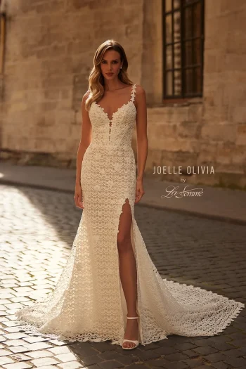A woman in the J2282 by Joelle Olivia fitted lace wedding dress, featuring a thigh-high slit, stands on a cobblestone street before a stone wall.