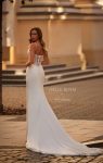 A woman in a fitted white J2275 by Joelle Olivia wedding dress with a lace bodice and thin straps stands outdoors in front of a classical building.