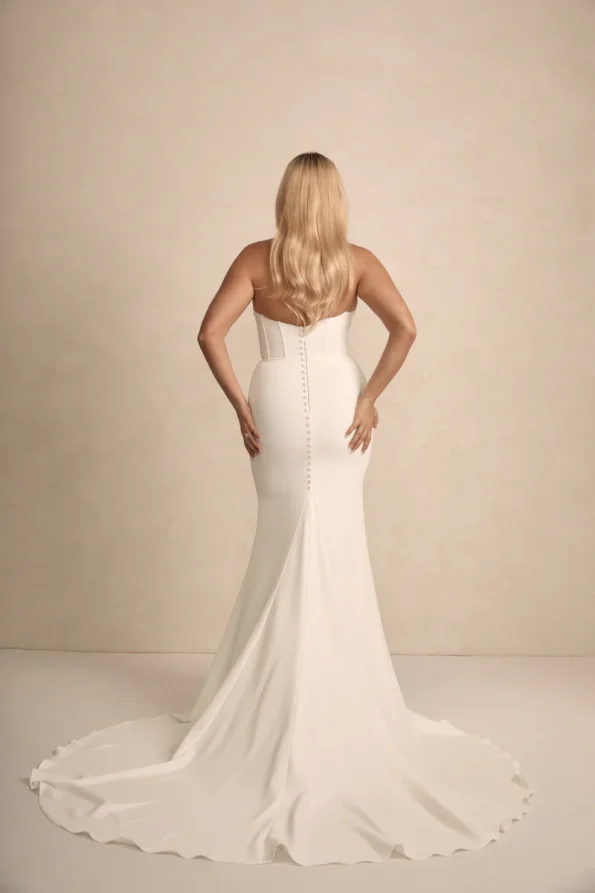 A woman with long blonde hair wears the strapless Rhode by Made with Love bridal gown, featuring a buttoned back and long train, standing against a plain light background.