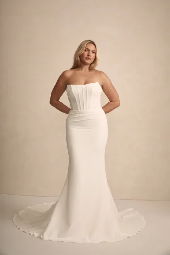 A woman stands before a plain background wearing the Rhode by Made with Love—a strapless, fitted white wedding gown featuring a structured bodice and a flared train.