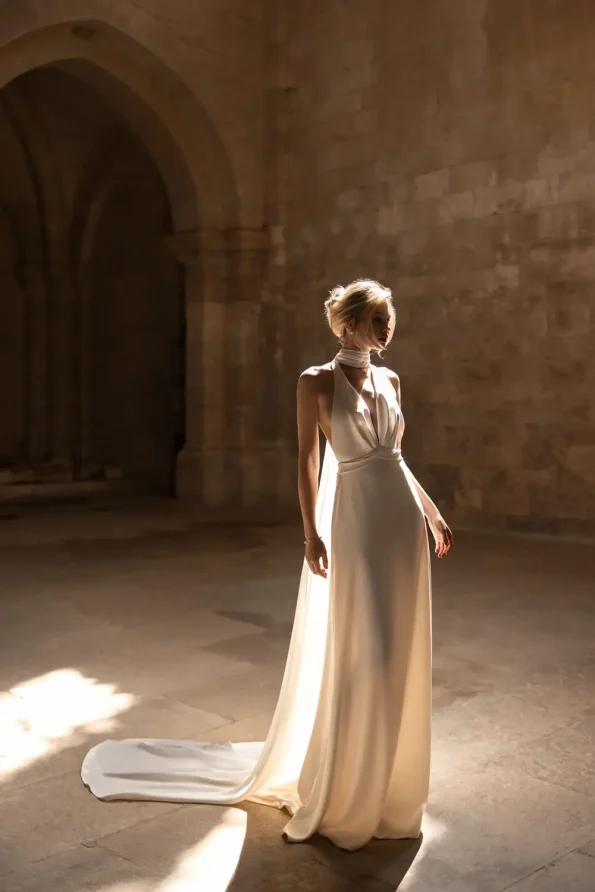 A woman in a flowing white Rouge by Eva Lendel halter-neck gown stands alone in a sunlit stone hall with arched doorways.