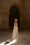 A woman in a flowing white Rouge by Eva Lendel halter-neck gown stands alone in a sunlit stone hall with arched doorways.